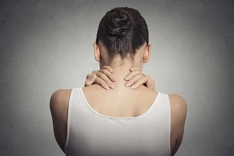 Frau in Rückenansicht hält sich vor Schmerzen mit leicht gesenkten Kopf den Nacken.