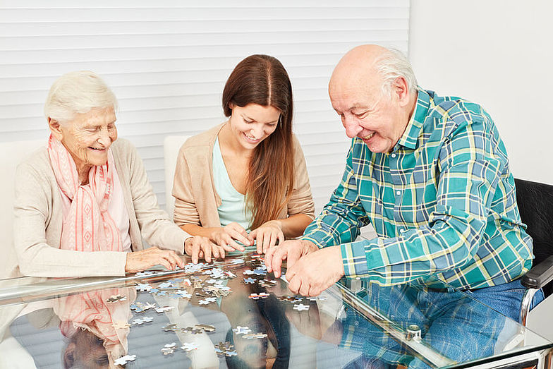 Eine ältere Damen und ein älterer Mann sitzen an einem Glastisch und puzzeln zusammen mit einer jungen Frau. Dabei sollen die Beweglichkeit der Finger und die geistigen Fähigkeiten trainiert werden. 