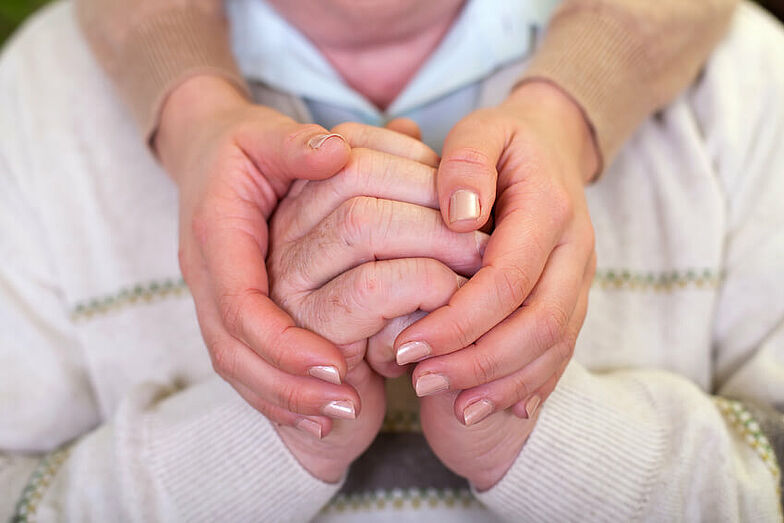 Ein Patient bekommt emotionalen Beistand, symbolisiert durch die Berührung der zusammengefalteten Hände durch eine andere Person.