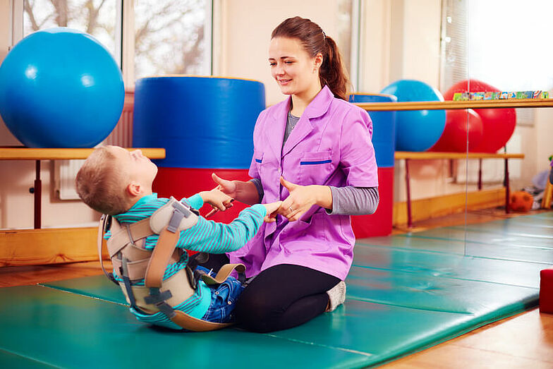 Ein kleiner Junge wird bei der Ausführung von Übungen von einer Therapeutin unterstützt.