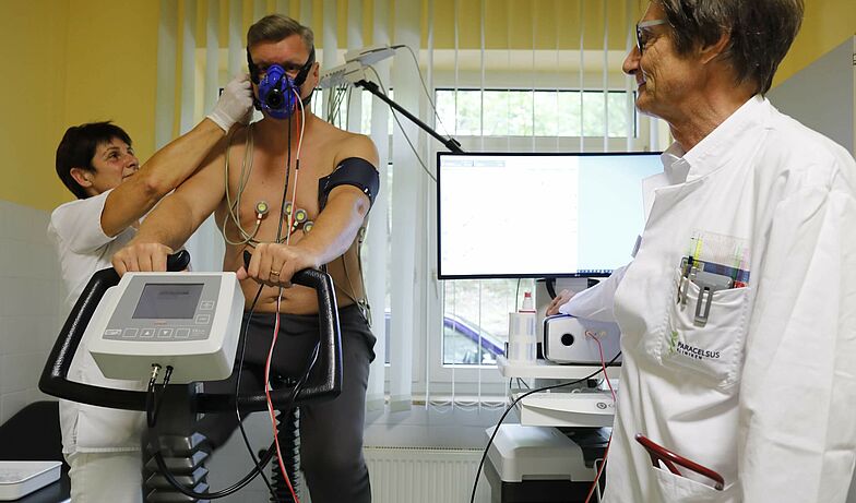 Lungenfunktionsdiagnostik bei Long Covid in Rehaklinik: Mann auf Fahrrad mit Atemmaske, Test wird beobachtet von Arzt und Pflegerin.