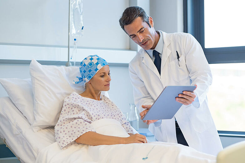 Eine Patientin mit Kopfbedeckung liegt in einem Krankenhausbett, schaut zusammen mit einem Arzt Unterlagen an, welche für die Chemotherapie relevant sind. 