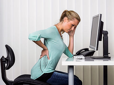 Bandscheibenvorfall: Frau mit Rückenschmerzen sitzt am Schreibtisch vor einem Computer.