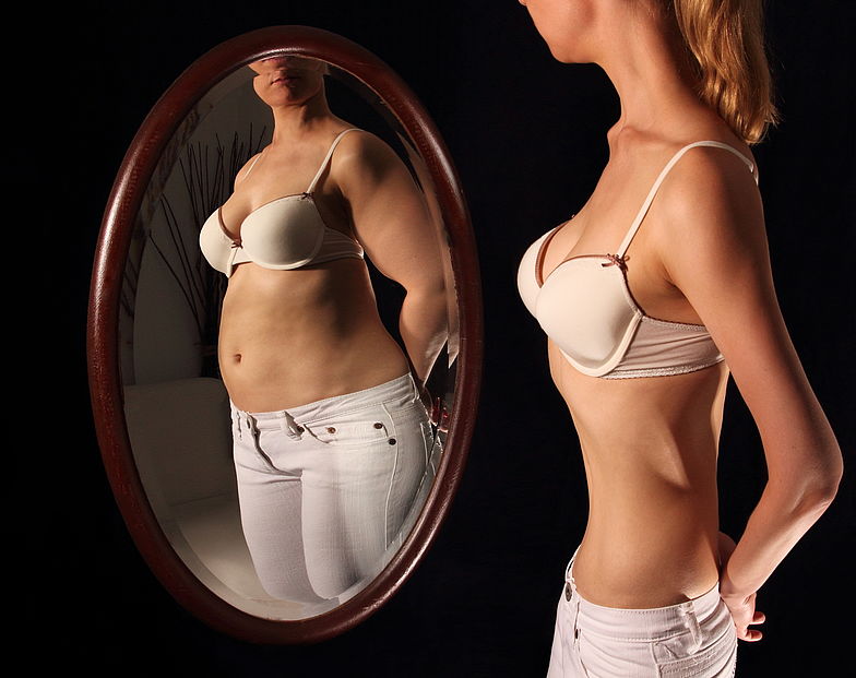 An einer Magersucht erkrankte Frau betrachtet sich im Spiegel. Das Spiegelbild zeigt die Frau mit Übergewicht.