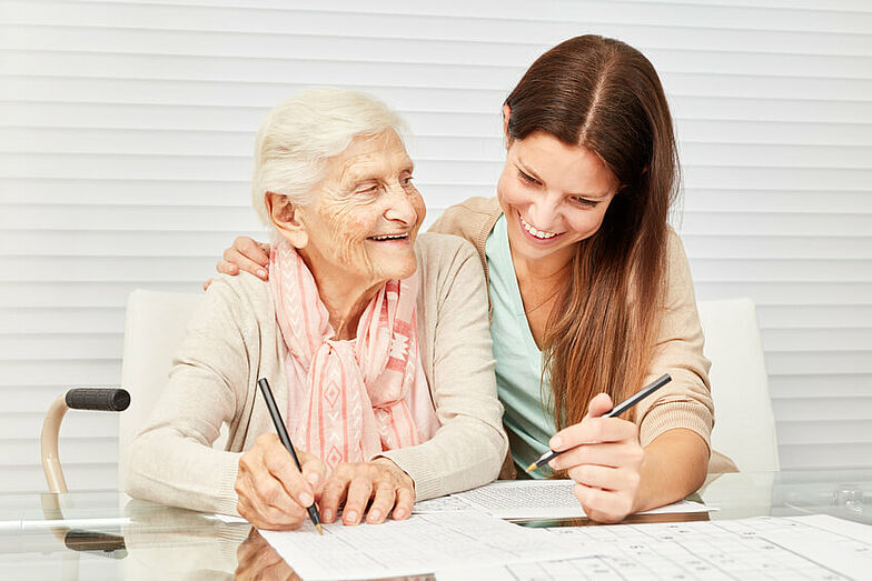 Eine ältere Dame wird durch eine Mitarbeiterin einer Rehaklinik bei dem Ausfüllen eines Sudokus unterstützt. 