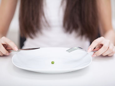 Reha bei Essstörungen: Frau sitzt mit Besteck in der Hand vor einem Teller, auf dem nur eine Erbse liegt.