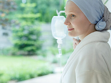 Eine Frau mit Kopfbedeckung berührt einen Infusionsständer, blickt aus dem Fenster in die grüne Natur und erholt sich von Chemotherapie.