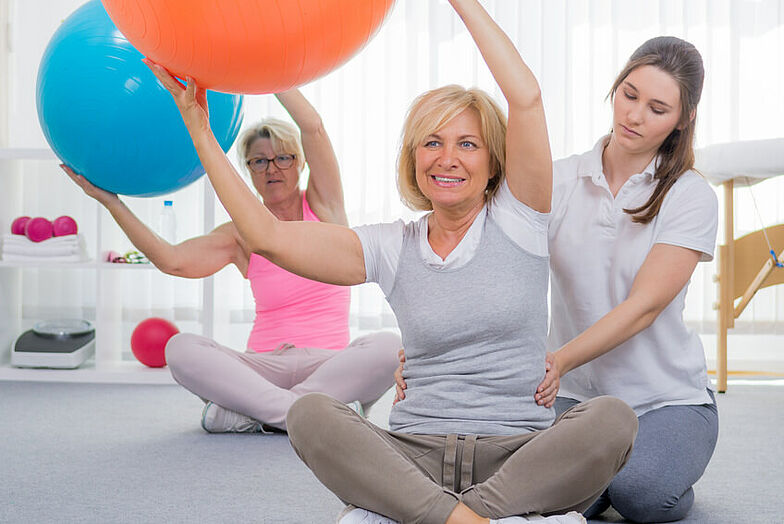 Zwei Frauen führen Übungen mit einem Gymnastikball aus. Dabei werden sie von einer Therapeutin unterstützt.