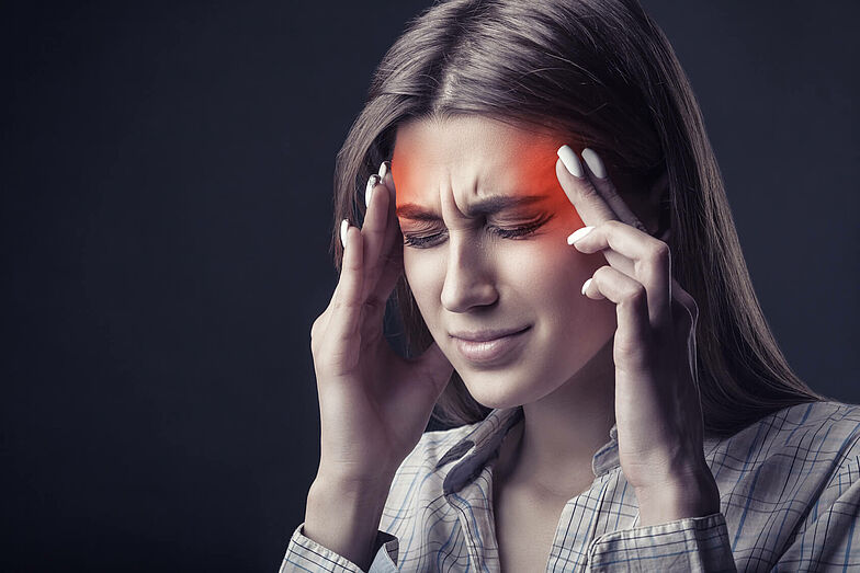 Eine Frau ist von Kopfschmerzen geplagt und fasst sich an die Schläfen. Der schmerzende Bereich ist rot eingefärbt.