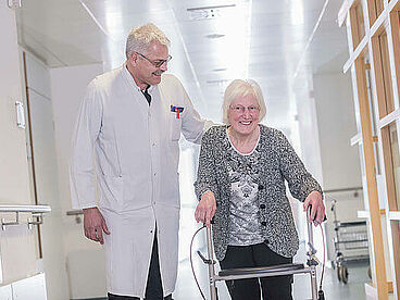 Arzt begleitet eine Patientin mit Rollator über den Klinikflur.